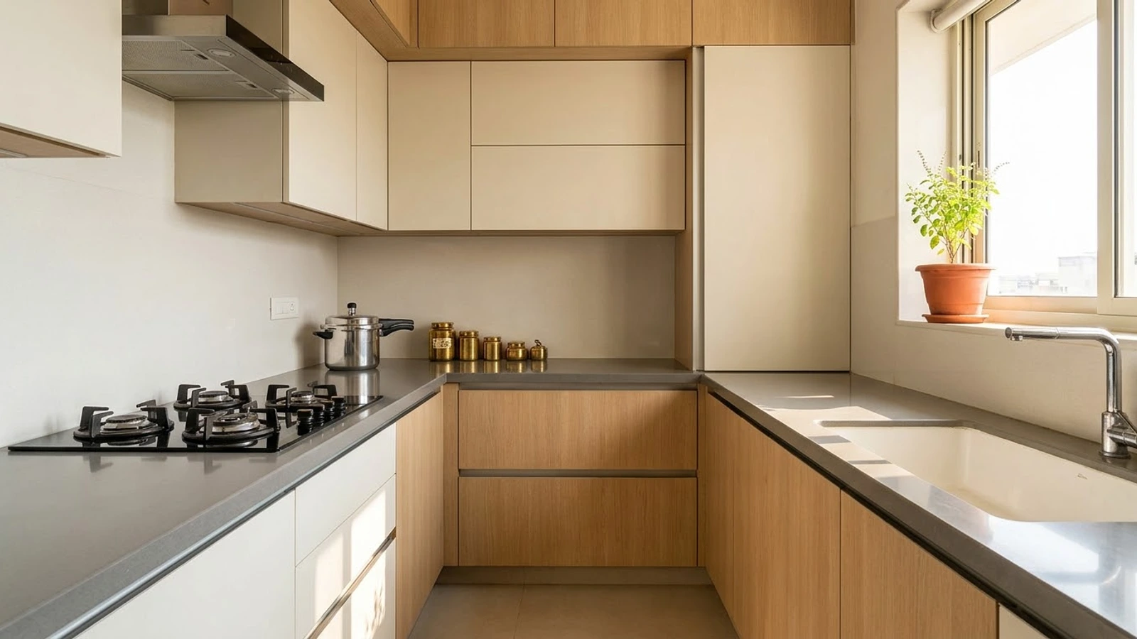 A modular kitchen in light wood and off-white with a gas hob on one counter and the sink on another by the window, a pressure cooker, brass jars and a tulsi plant on the sill