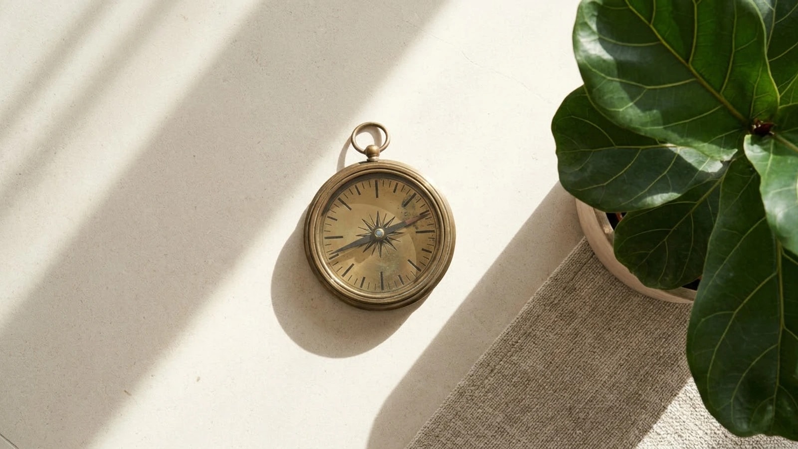 A brass compass on a pale stone floor crossed by bands of morning sunlight, beside a linen rug and a potted plant