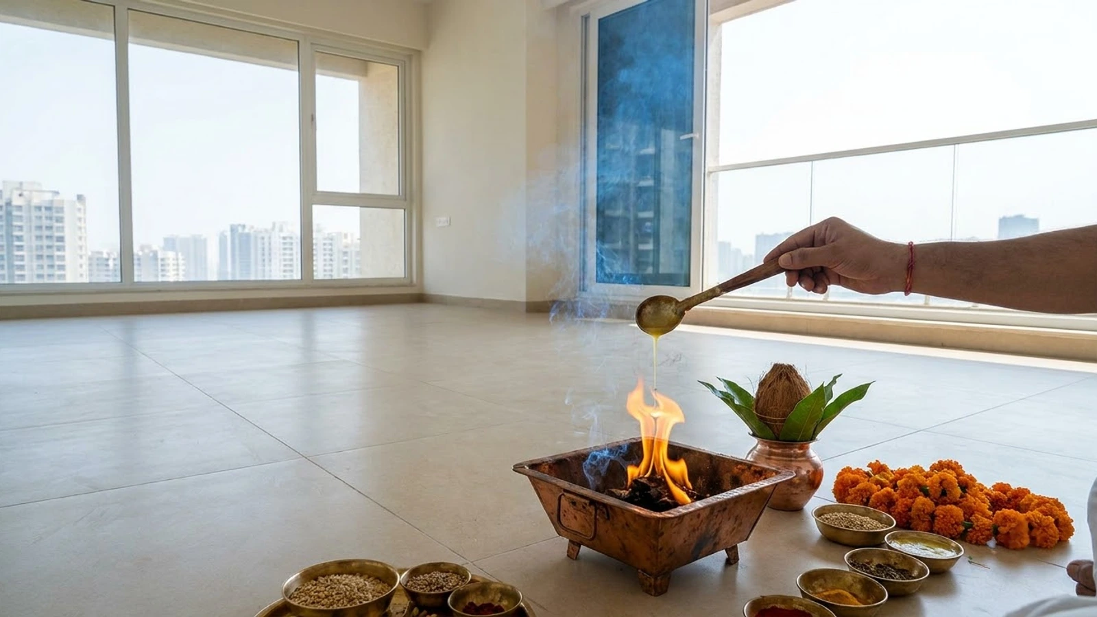 A hand pouring ghee from a long spoon into the fire of a copper havan kund on the floor of an empty new flat, beside a kalash topped with mango leaves and a coconut, marigolds and bowls of offerings