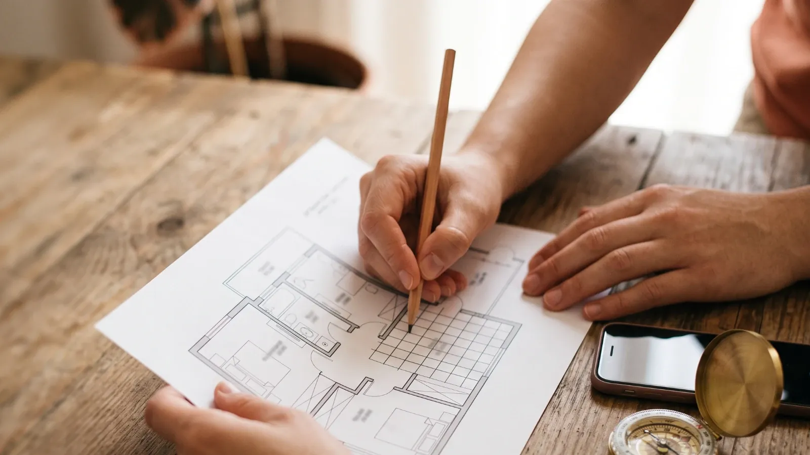 Hands drawing a pencil grid over a printed floor plan on a wooden table, with a phone and a brass compass beside it