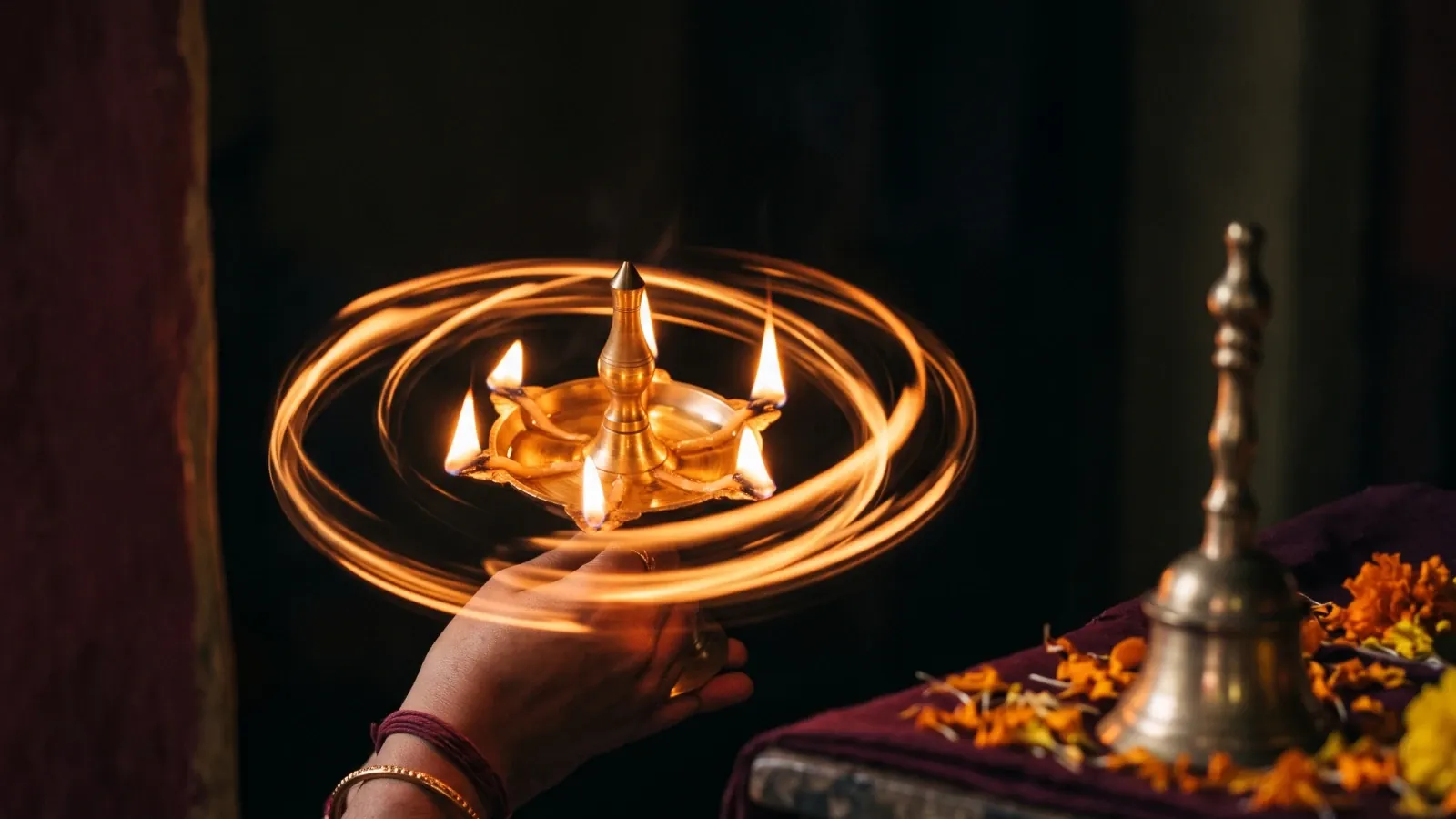 A hand circling a five-wick brass aarti lamp, its flames leaving rings of light in a dark room, beside a brass bell and marigold petals