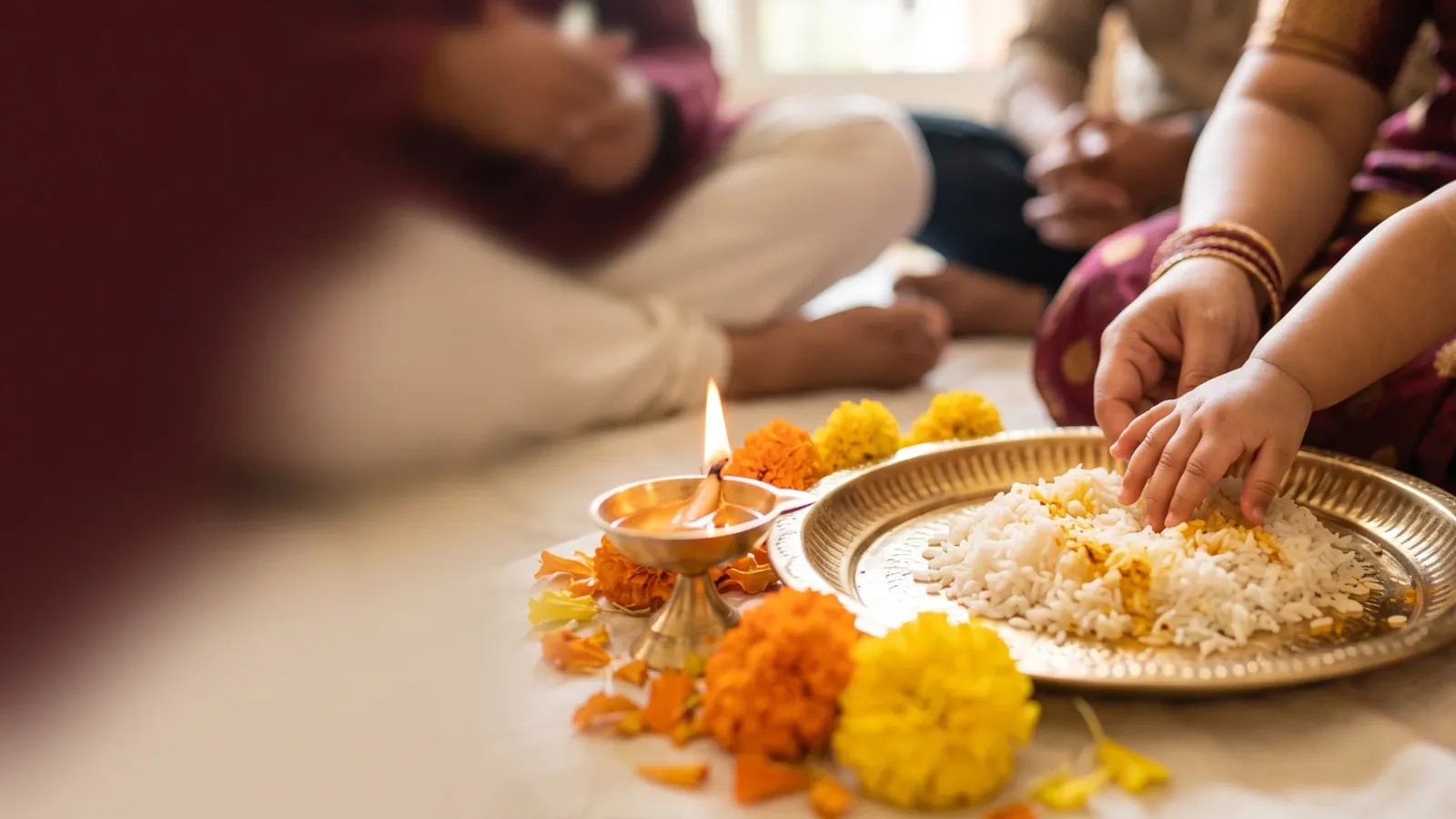 A baby’s small hand reaching into a brass plate of rice beside a lit diya and marigolds, with family members seated around