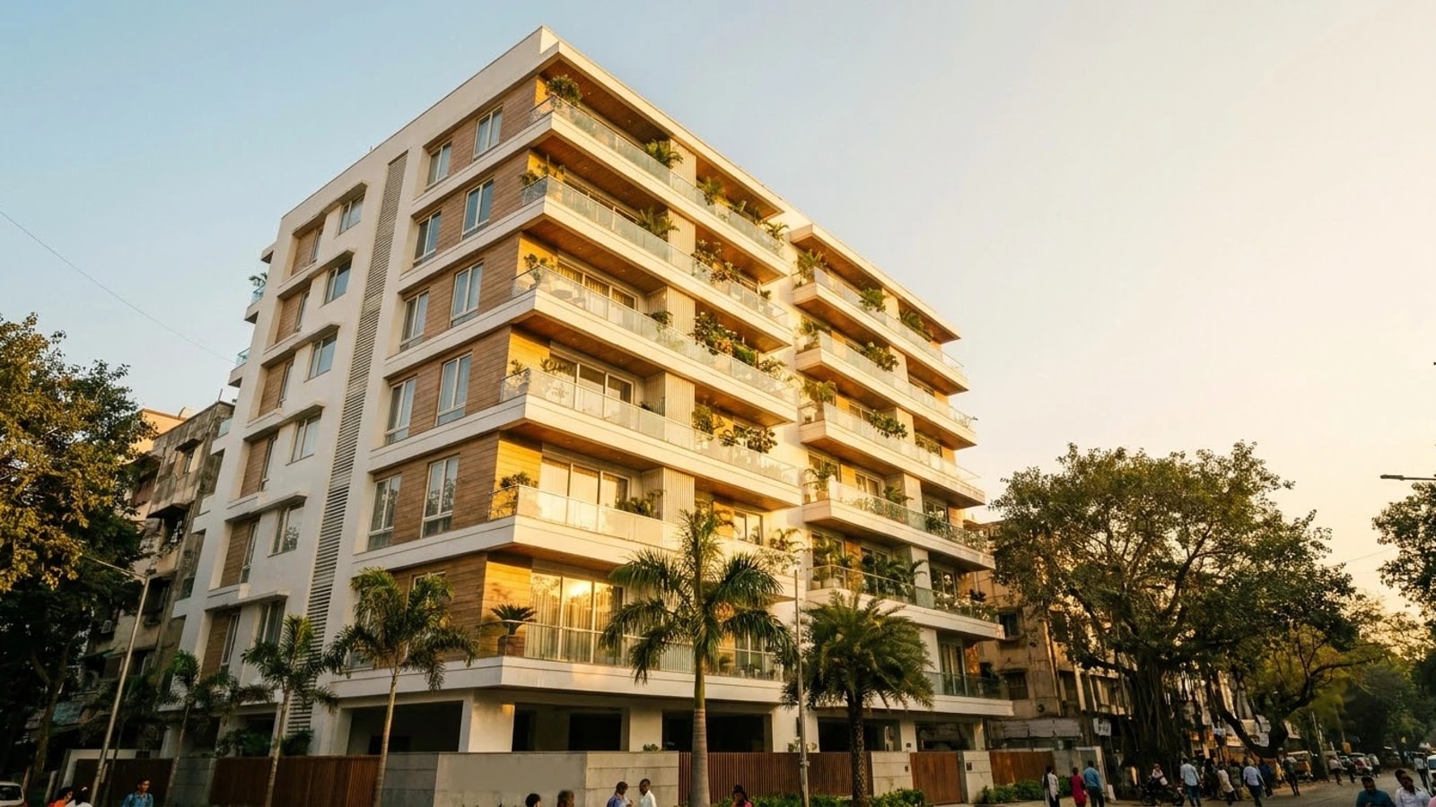 A modern apartment building in an Indian city at golden hour, seen from the street, with glass-railed balconies full of plants