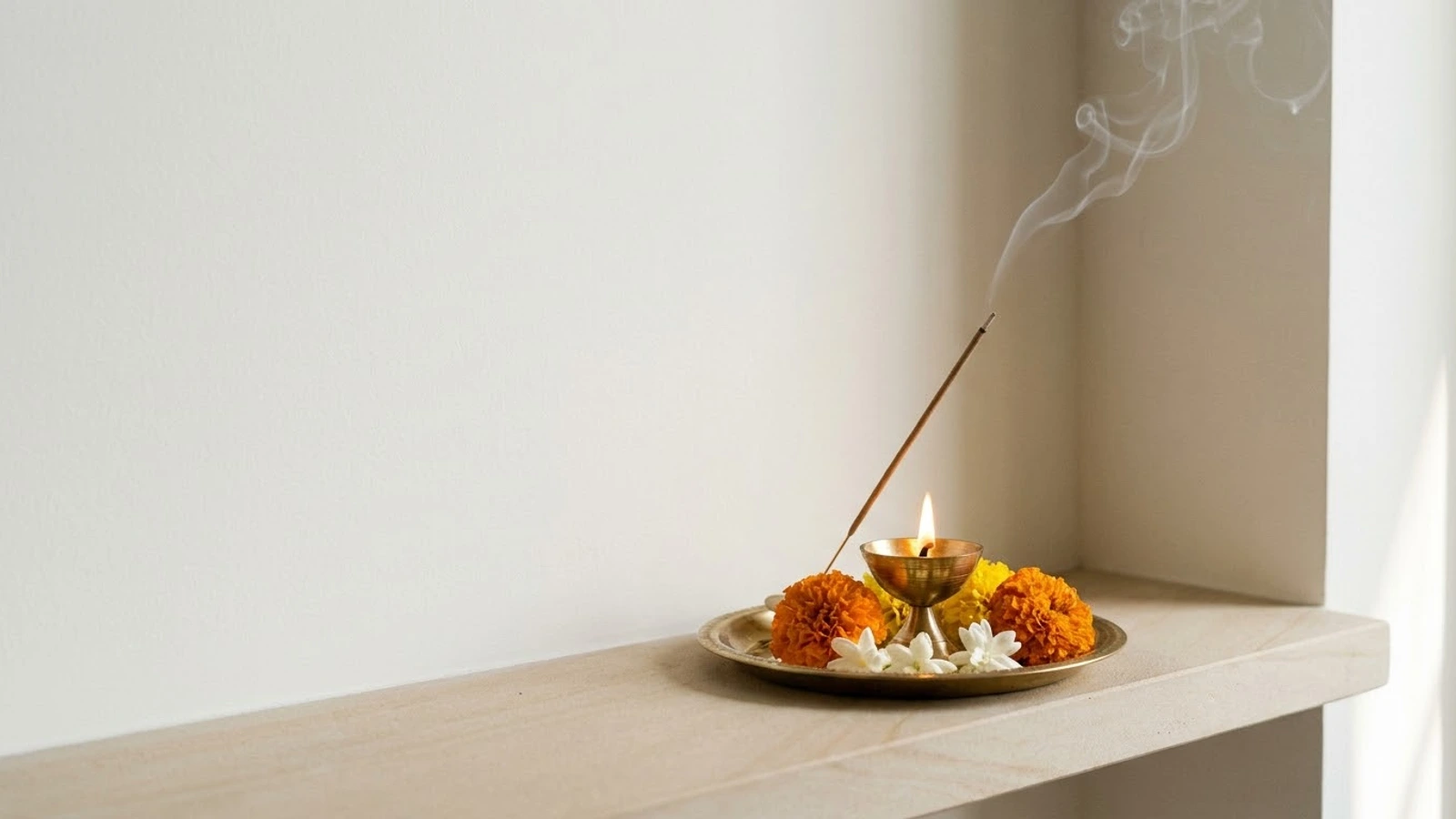 A lit brass diya on a brass plate with marigold and jasmine flowers and a stick of incense, its smoke rising against a plain white wall